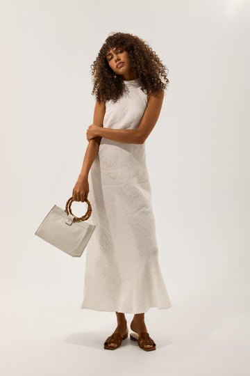 Woman in a white sleeveless linen maxi dress carrying a bamboo handle handbag with brown leather sandals.
